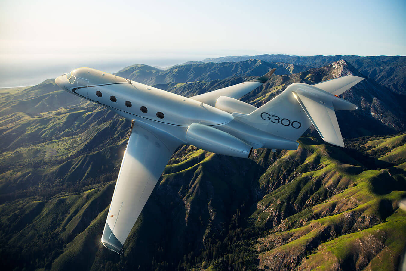A Gulfstream G300 flies above mountainous, forested terrain.