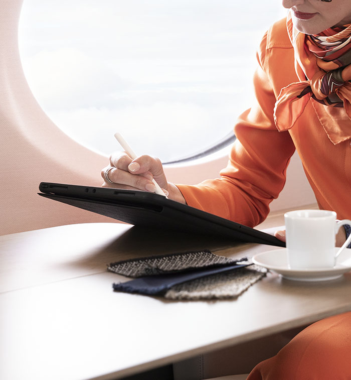 A fashion executive sitting next to a large oval window works as her tablet computer and coffee rest on an extended work surface.