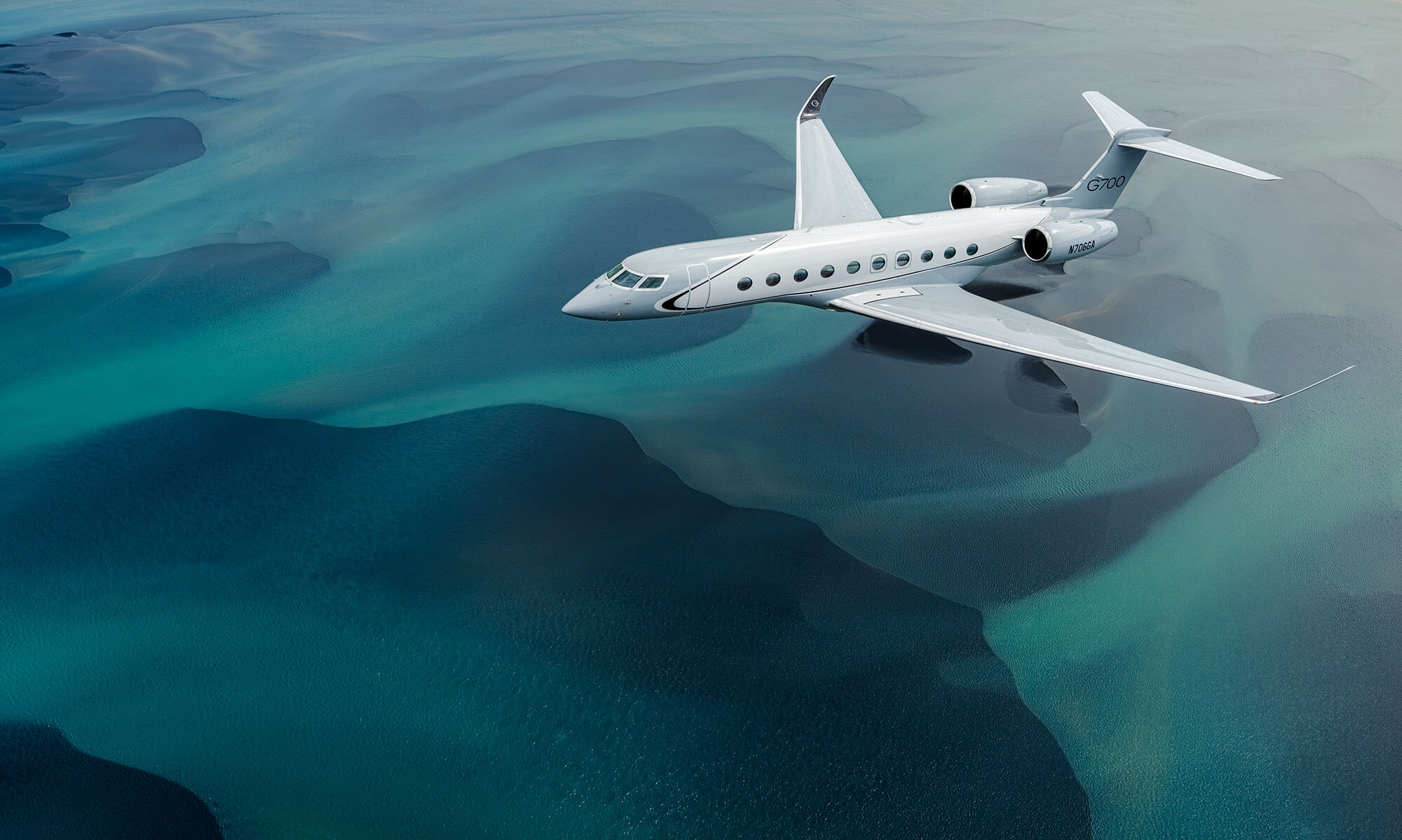 A Gulfstream G700 aircraft seen from slightly above and behind flies over rocky, snow-covered mountain peaks.