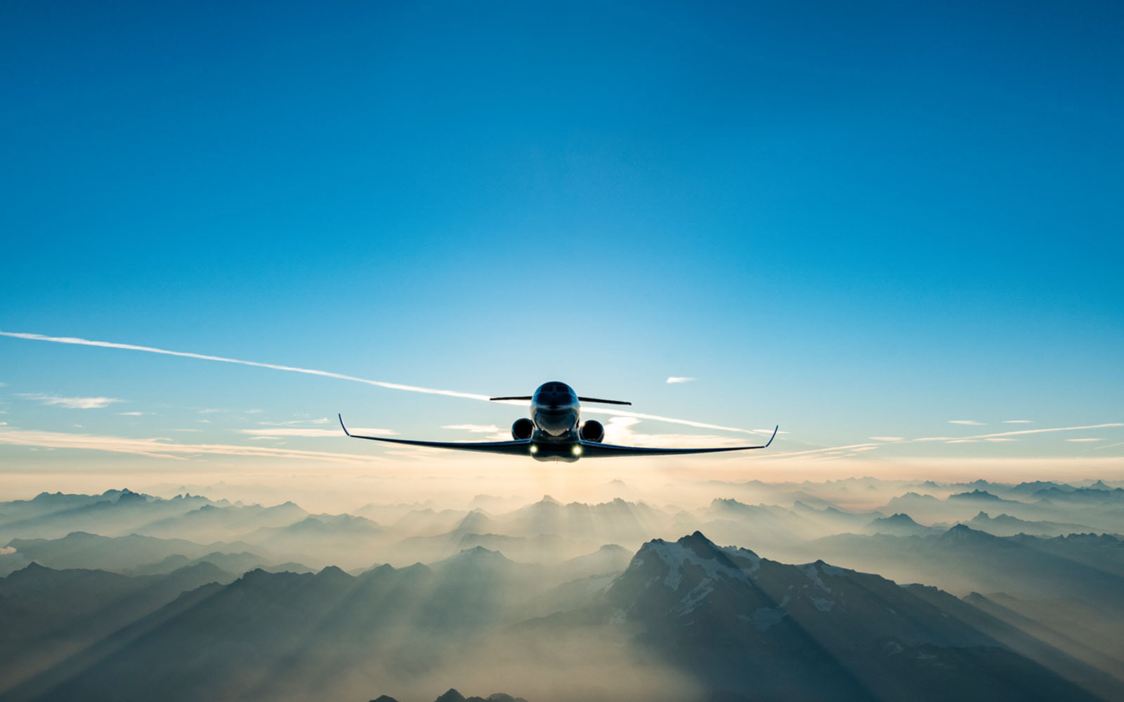 A Gulfstream private aircraft flying toward viewer above misty mountain peaks and backlit by morning sun