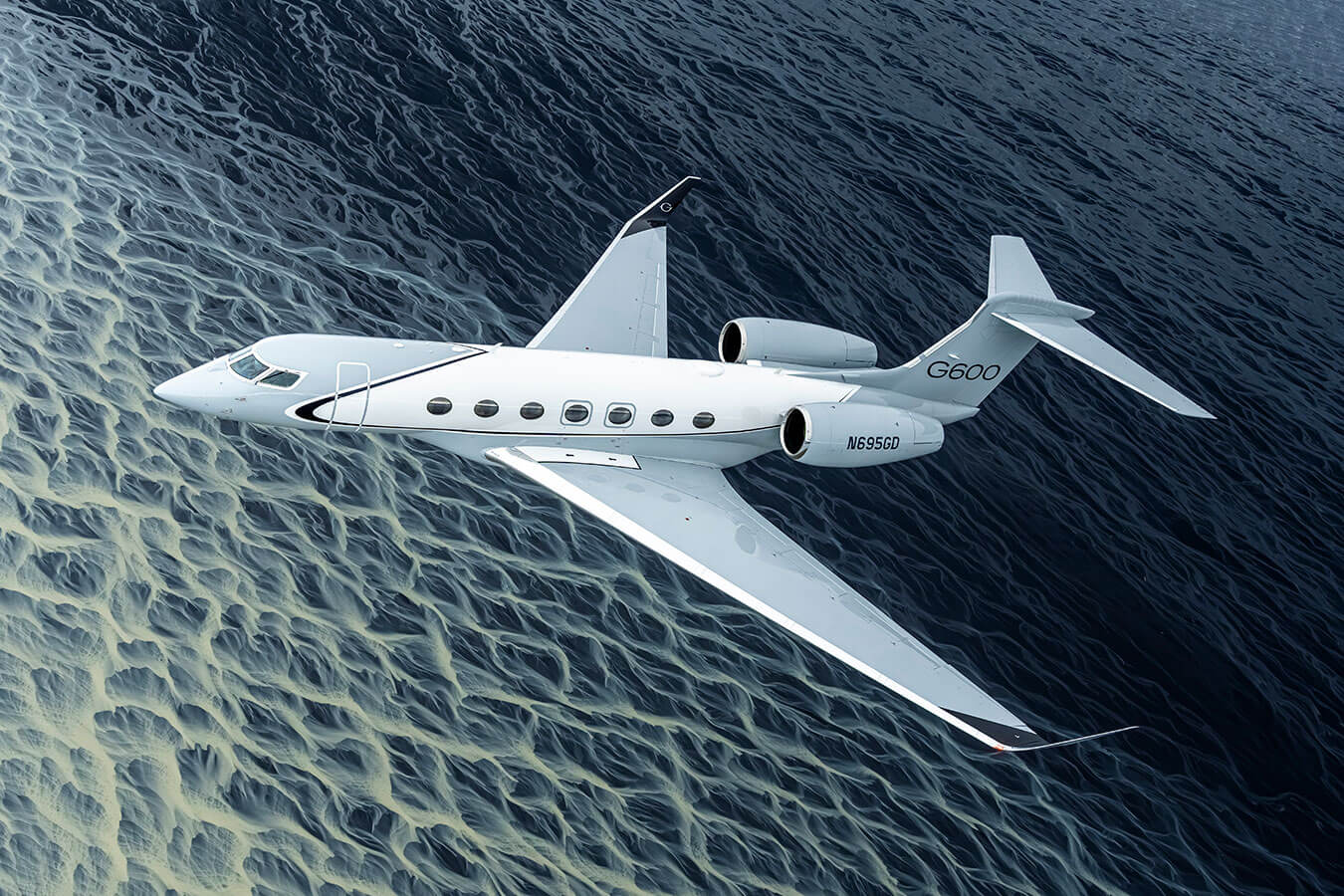 A Gulfstream G600 flies above blue water with whitecaps criss-crossing in a geometric pattern.