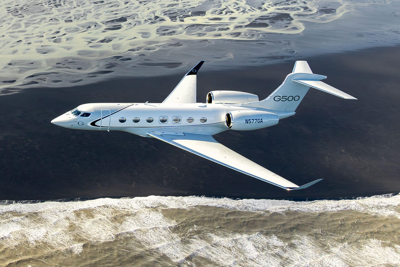 A Gulfstream G500 flies above snow-dusted brown dunes lapped by the waves of a shallow coastal channel.