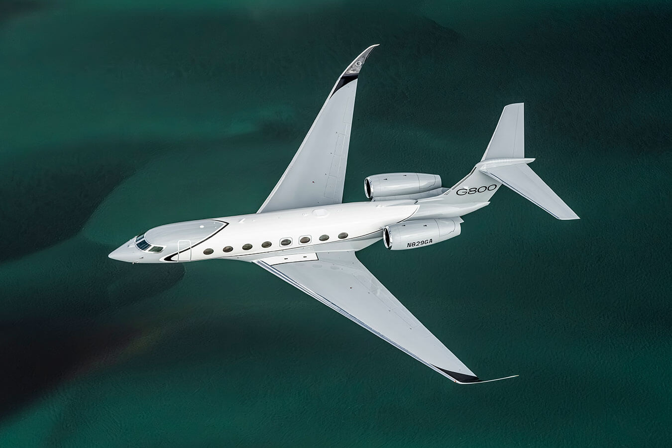 A Gulfstream G800 flies over shallow emerald green water.